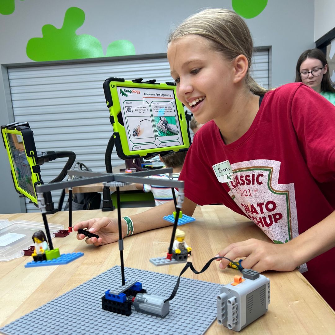 A child smiling while building a LEGO robotics project at a classroom table, following step-by-step instructions on a tablet during a hands-on STEM activity.